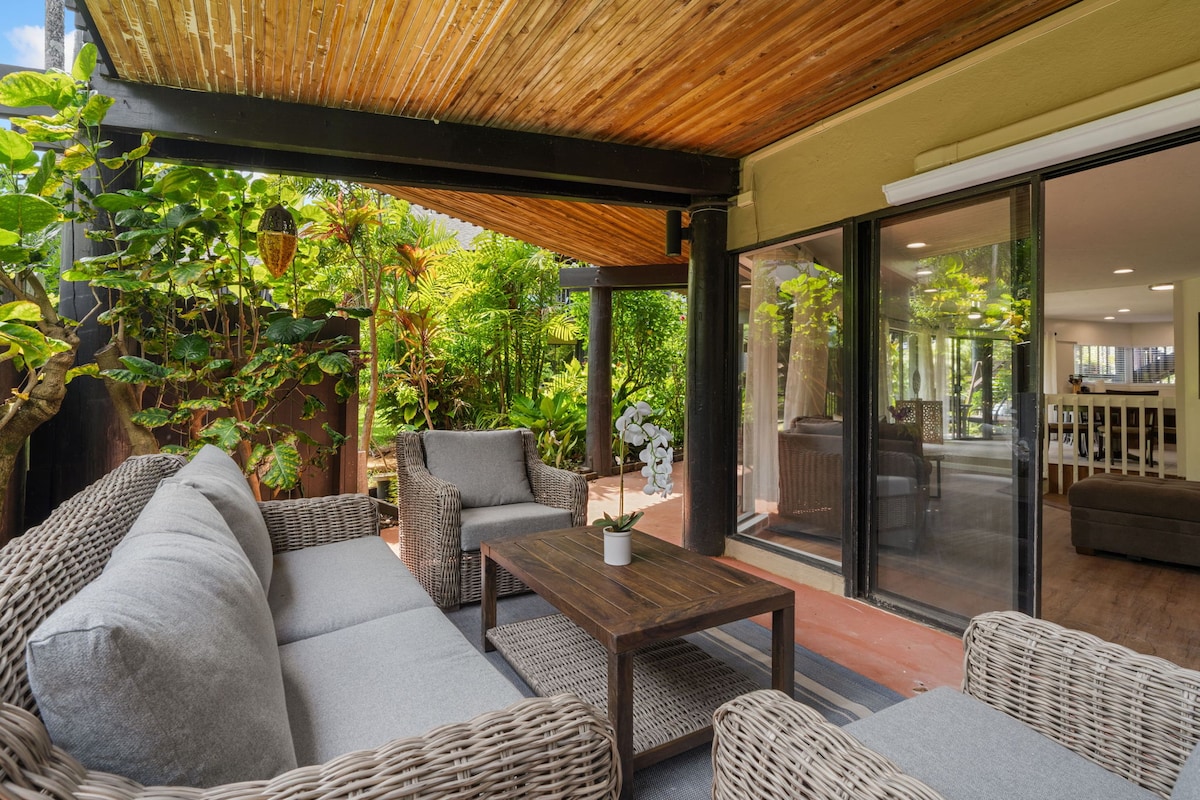 A covered outdoor seating area is furnished with comfortable, gray wicker sofas surrounding a wooden coffee table. Lush greenery provides a natural backdrop, enhancing the serene atmosphere. Large windows allow for a seamless transition between indoor and outdoor living spaces.