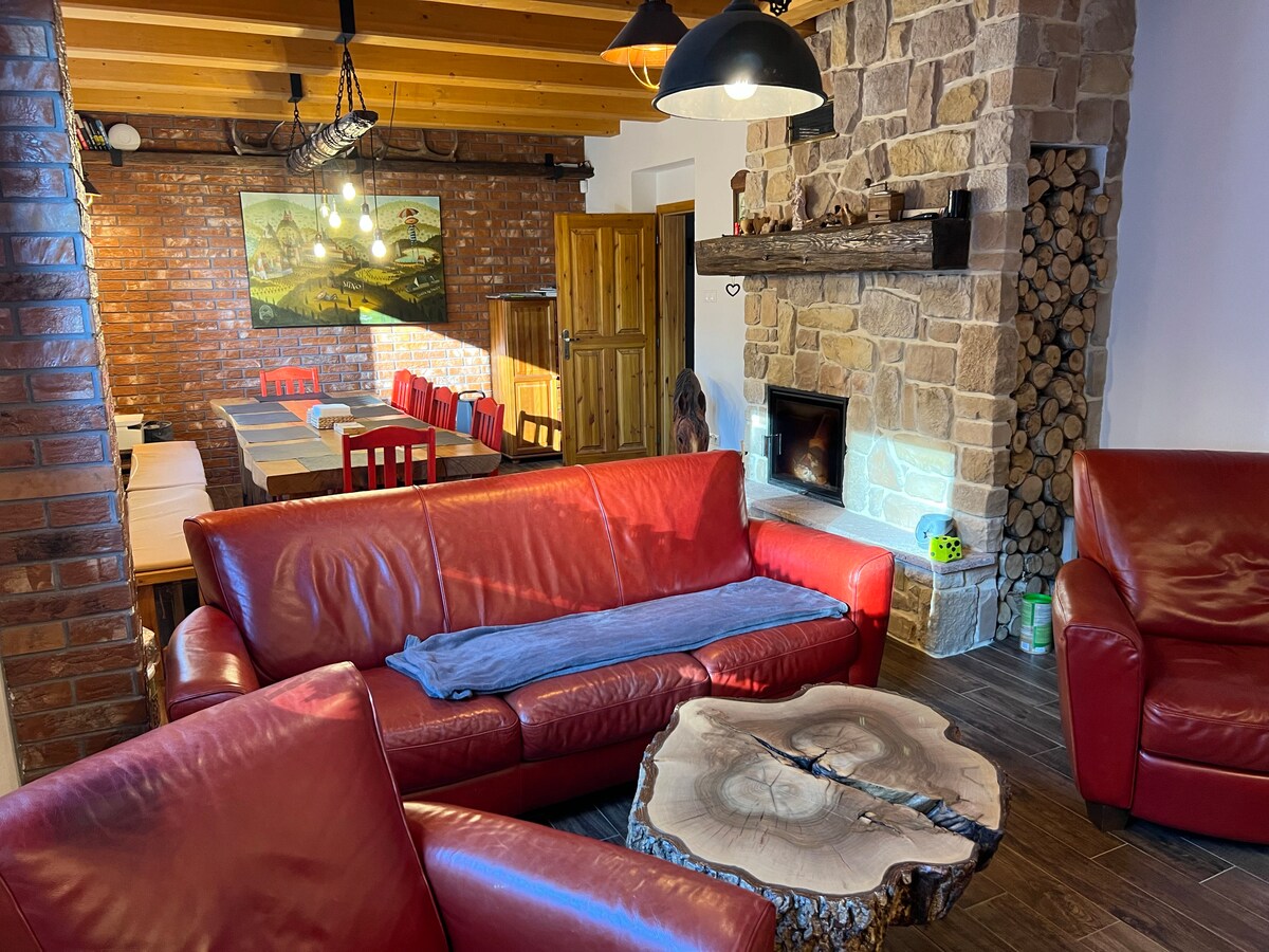 A cozy common room is showcased with red leather sofas positioned around a unique wooden table. A stone fireplace adds warmth, while a large artwork of a rustic landscape adorns the wall. The dining area is visible in the background, featuring a lengthy table and red chairs.