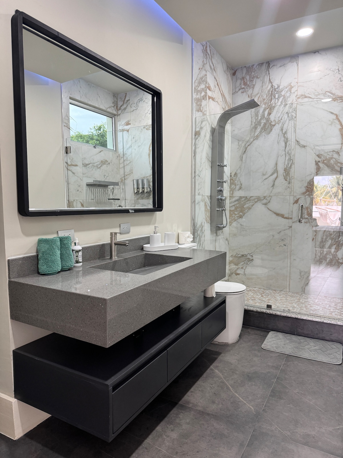 A modern bathroom features a sleek countertop with a large mirror above. The gray stone sink is complemented by neatly arranged toiletries. A glass shower enclosure showcases marble-patterned walls, while a subtle blue light adds a soft glow to the space. A small rug lies on the floor.