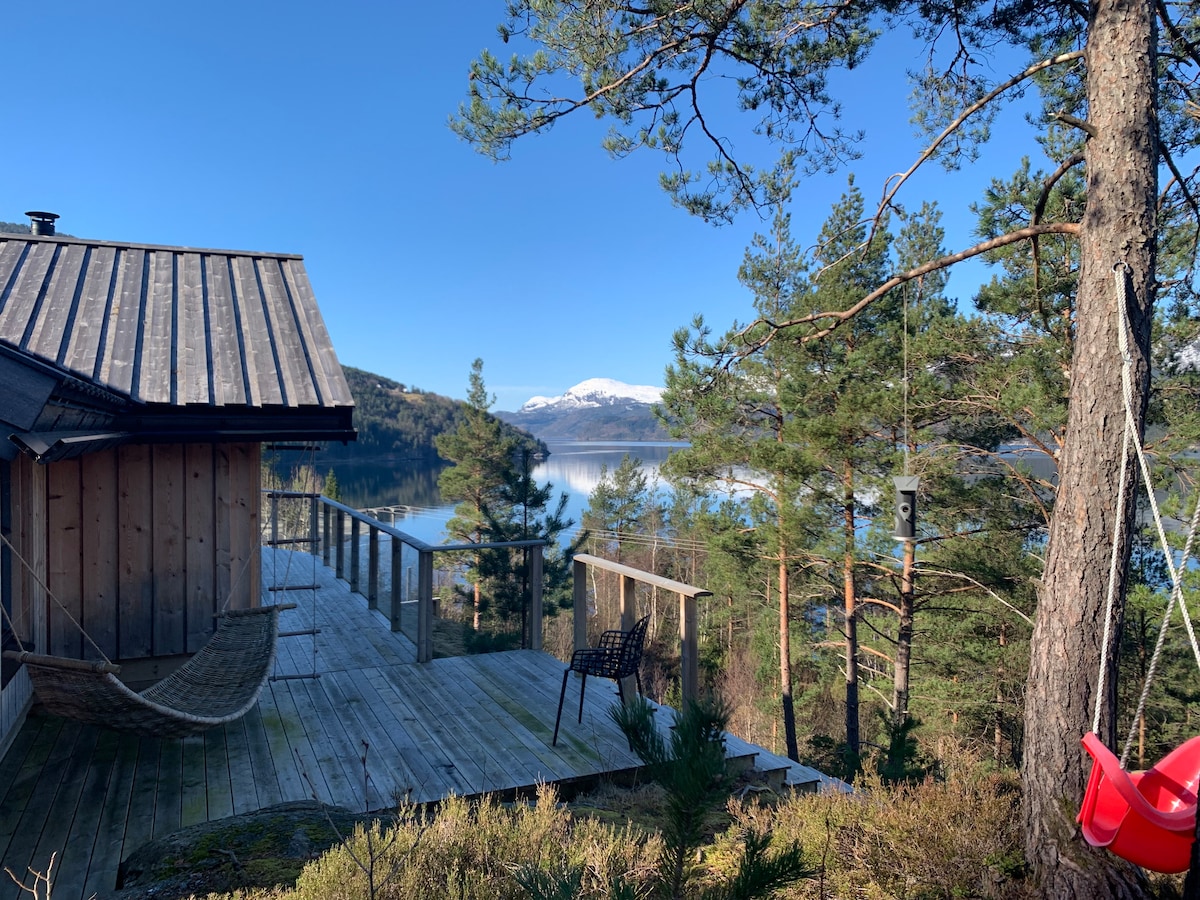 A wooden deck extends from the cabin, offering unobstructed views of the fjord and surrounding mountains. Pine trees frame the scene, and a black chair is positioned near the edge. The clear blue sky reflects on the calm water below.