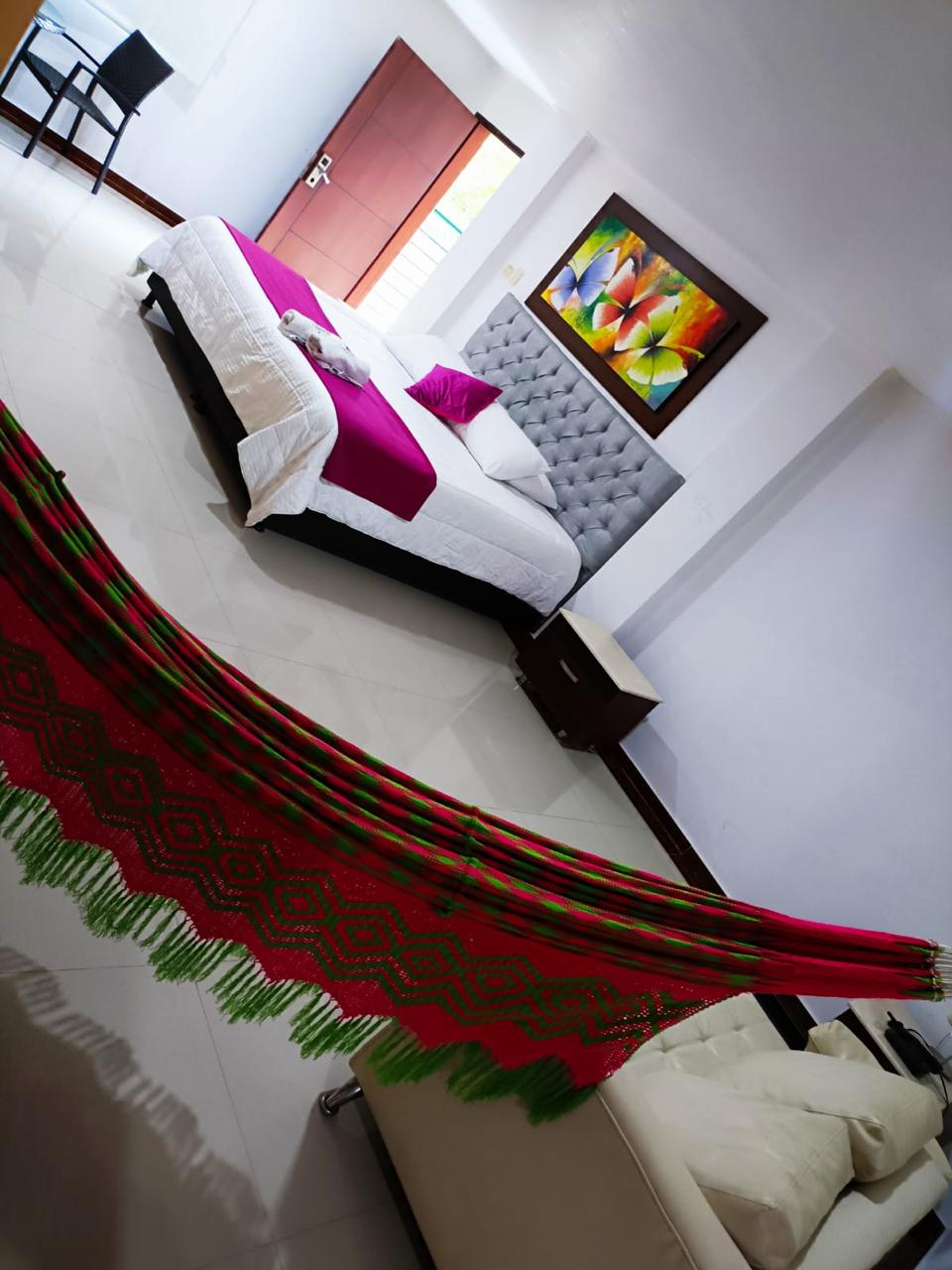 A spacious room features a large bed with a plush headboard and colorful accents. A vibrant hammock hangs in the center, drawing attention. Art is displayed on the wall, and ample light comes through the windows, illuminating the modern furnishings and bright decor.