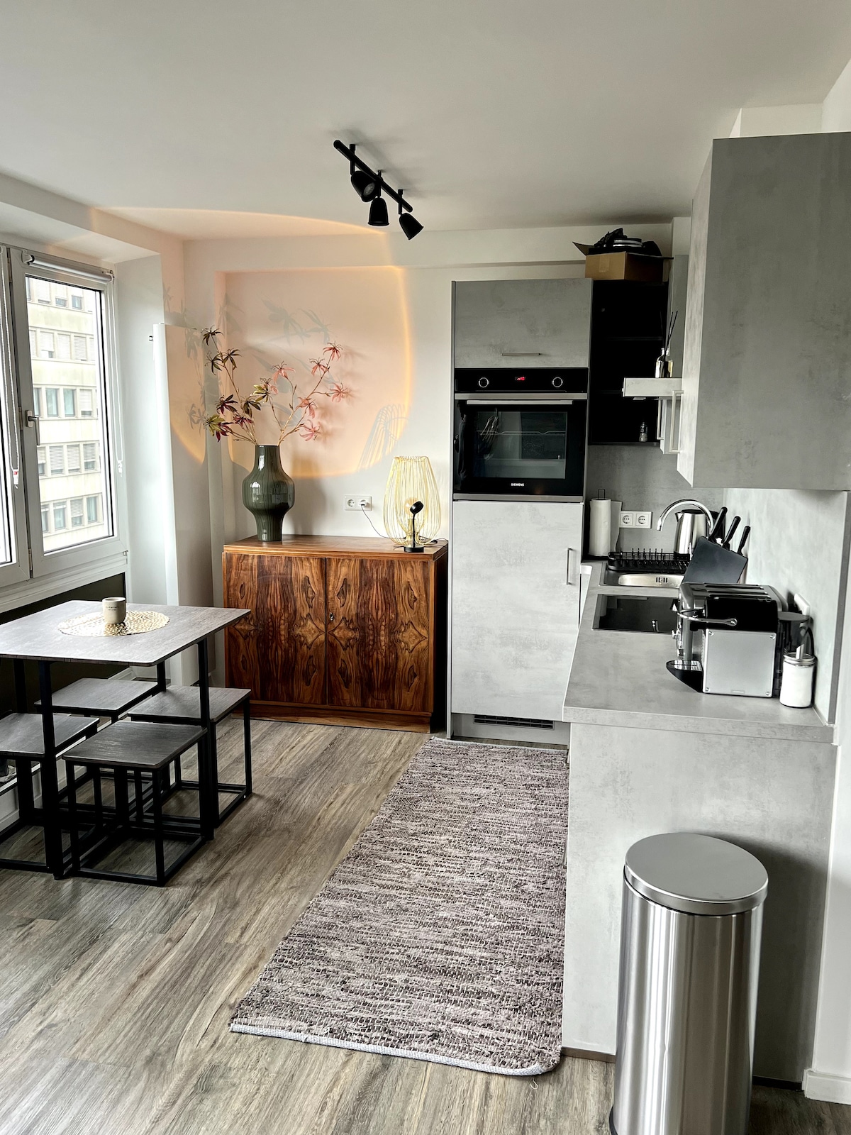 A modern kitchen features sleek gray cabinetry and a built-in oven. A compact dining table is positioned beside large windows, allowing natural light to brighten the space. Decorative elements include a vase with foliage on the countertop and a textured area rug on the floor.