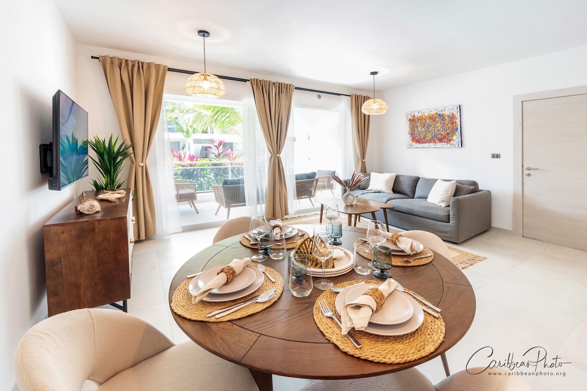 A spacious dining area is showcased, featuring a round wooden table elegantly set for a meal. Comfortable chairs surround the table, and a cozy sofa is positioned nearby. Large sliding glass doors lead to a balcony with tropical views, complemented by soft lighting fixtures.