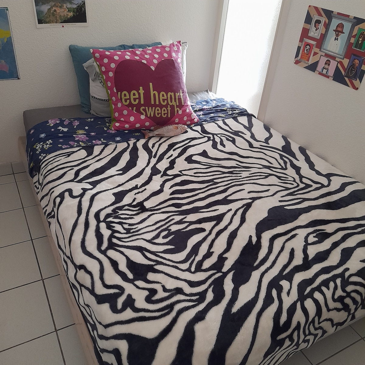 A bed features a patterned zebra-print blanket layered over a gray fitted sheet. Plush pillows, including one with a heart design, are arranged at the head of the bed. The room is brightened by natural light filtering through a nearby window.