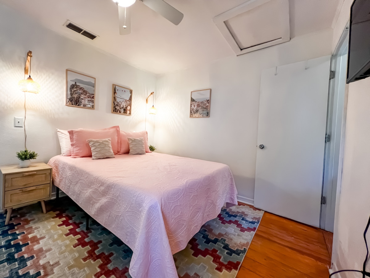 A bedroom with a queen bed features soft pink bedding and decorative pillows. Wall lights illuminate the space beside framed artwork. A small nightstand holds a plant, while a colorful area rug adds warmth. A flat-screen TV is mounted on the opposite wall.