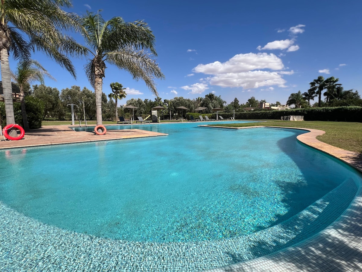 A large pool is surrounded by manicured lawns and palm trees, reflecting the clear blue sky. Lounge chairs line one side, and a gentle slope leads to the shallow end, providing a serene setting for relaxation.