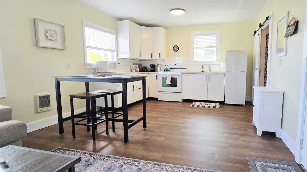 The kitchen area features modern cabinetry and a small dining table with two stools. Natural light is provided by two windows above the sink. Stainless steel appliances, including a refrigerator and stove, are complemented by light-colored walls and wood flooring.