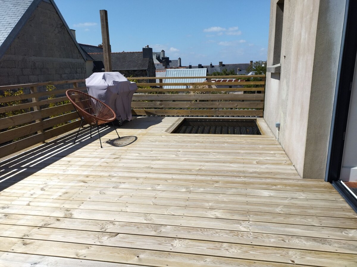 A spacious wooden terrace features a built-in grill, surrounded by a wooden railing. A round, woven chair is positioned nearby, while a sunken area on the floor hints at further outdoor seating. Clear blue skies complement the tranquil atmosphere.