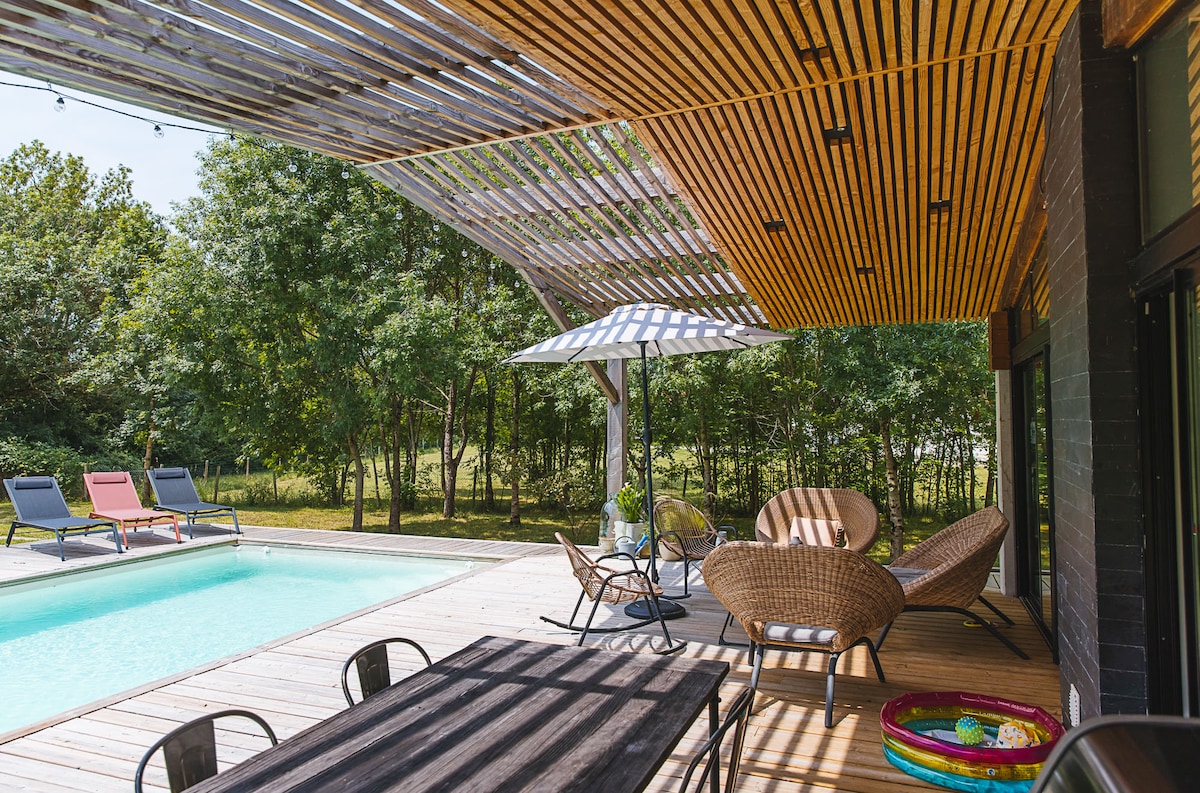 An outdoor terrace is featured, equipped with a spacious dining table and a variety of seating options. A well-maintained pool is visible nearby, framed by shaded areas from a wooden canopy and a large umbrella. Lush greenery surrounds the space, enhancing its tranquil setting.