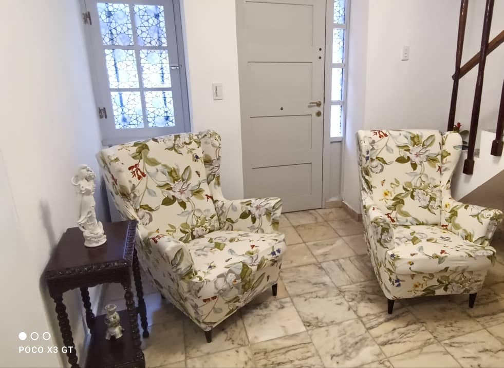 A small sitting area features two floral patterned armchairs positioned beside a wooden side table. Light-colored marble flooring enhances the space, while a decorative figurine is placed on the table. A door with glass panels provides a view of the exterior.