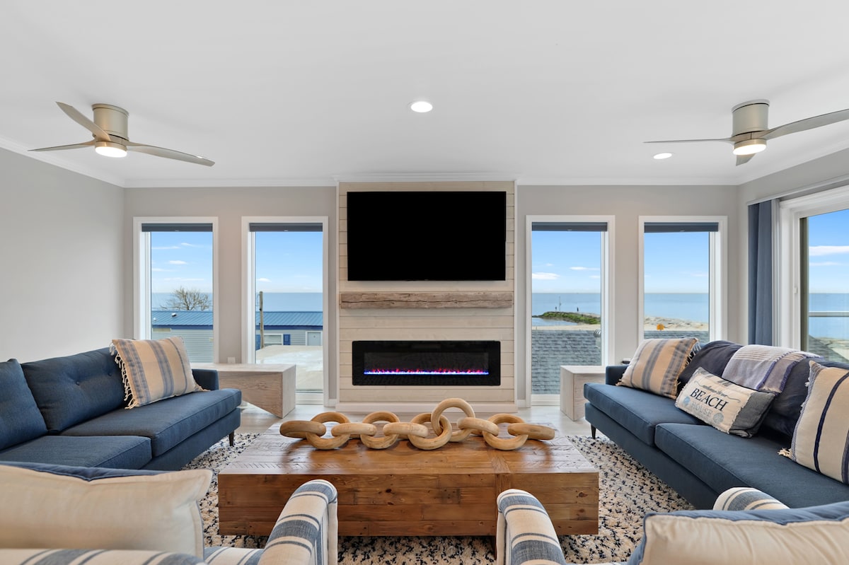 The living area features two comfortable sofas facing a modern fireplace and a large wall-mounted television. Floor-to-ceiling windows provide views of the Delaware Bay. A wooden coffee table with decorative accents sits in the center, complemented by a light-colored area rug beneath.