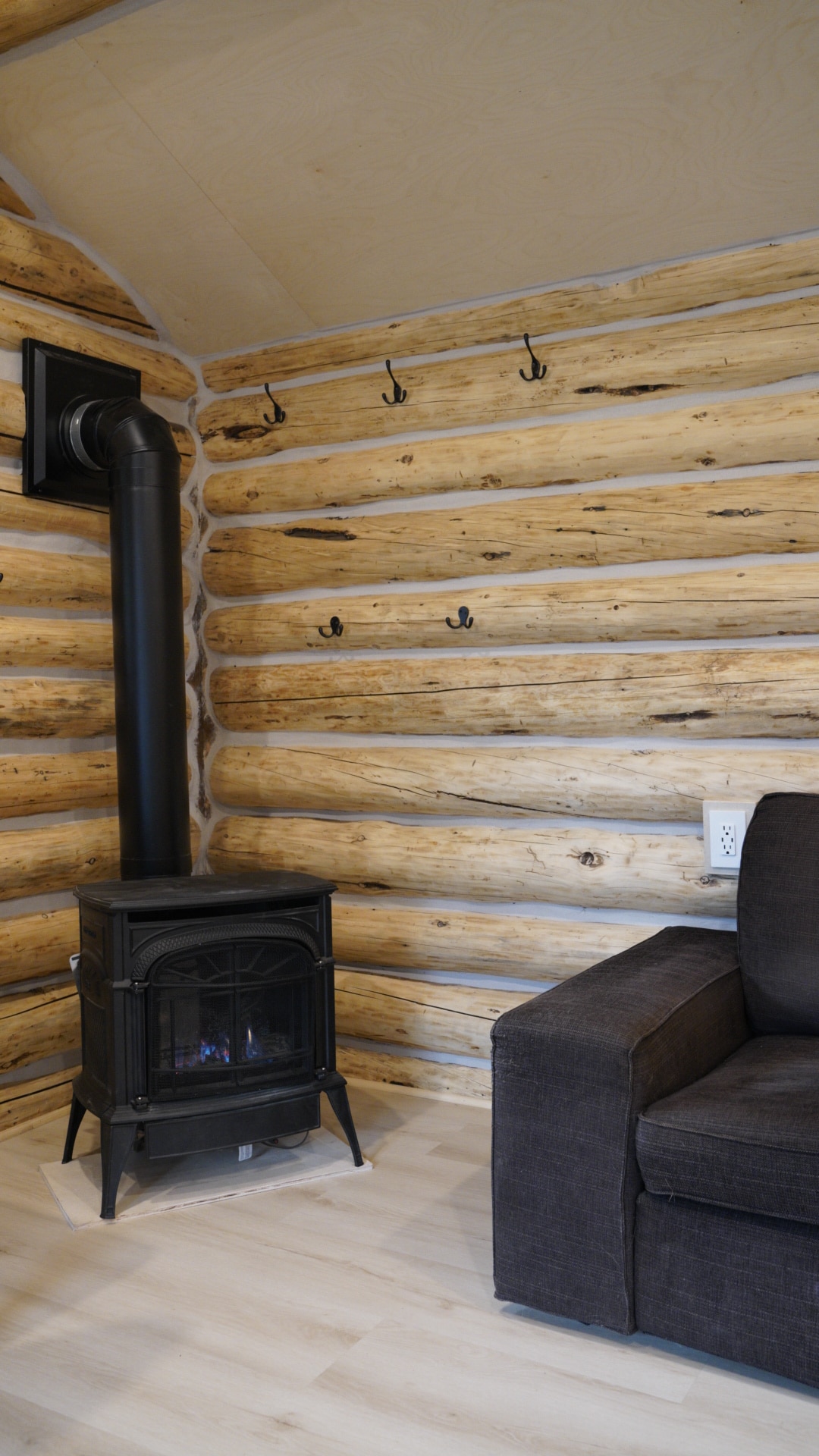 A cozy corner of the cabin features a black stove on a light wooden floor, surrounded by natural log walls. Above the stove, several hooks are mounted for hanging items. A dark sofa is positioned nearby, creating a comfortable setting.