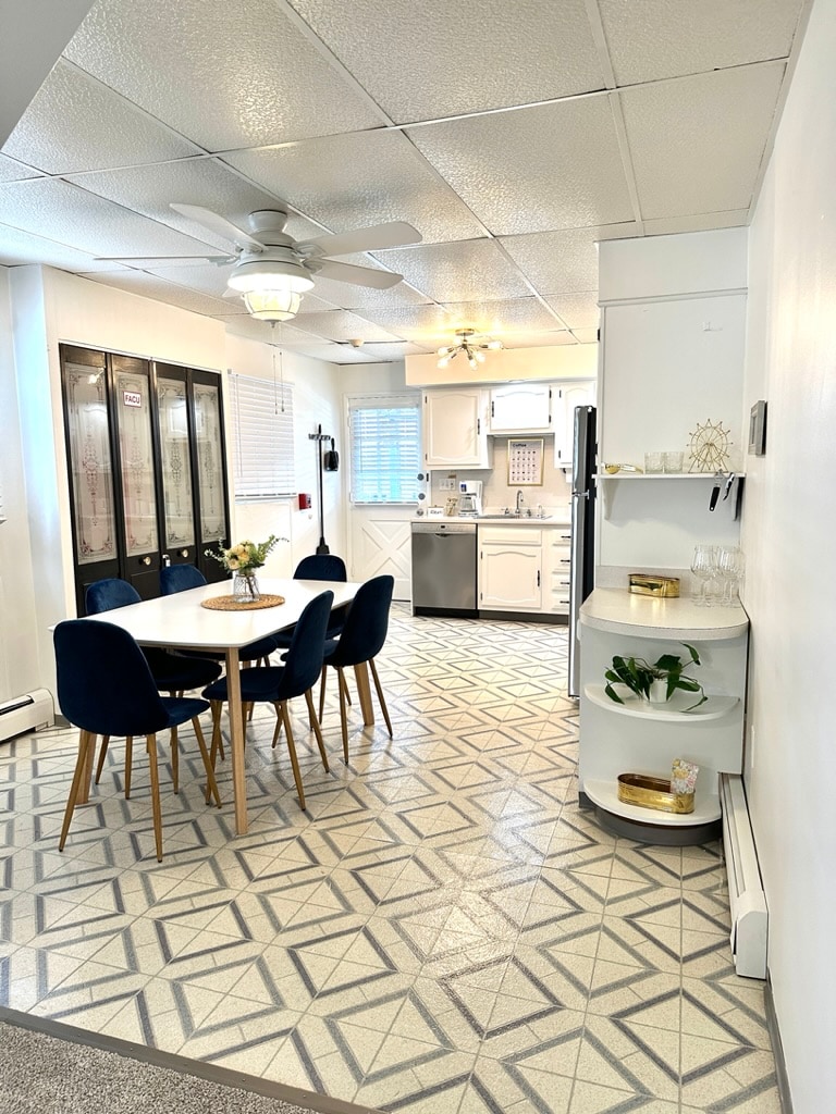 A spacious dining area is visible, featuring a round table surrounded by six navy upholstered chairs. The modern kitchen is equipped with stainless steel appliances and offers storage space. Natural light enters through the bright window, and a patterned floor adds a unique touch.