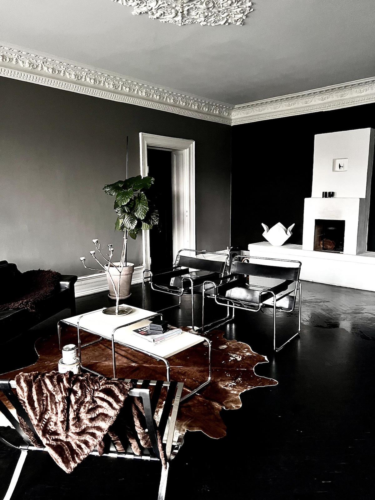 A spacious living room features a blend of modern and classic design elements. A comfortable black sofa is paired with stylish metallic chairs around a glass coffee table. A decorative fireplace is located on one wall, complemented by a potted plant and a unique cowhide rug on the floor.