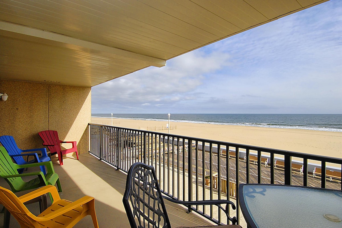 A spacious balcony offers a relaxing view of the sandy beach and ocean. Colorful Adirondack chairs in vibrant hues are positioned for comfort alongside a round table, creating an inviting outdoor space. The horizon meets the gently rolling waves under a partly cloudy sky.