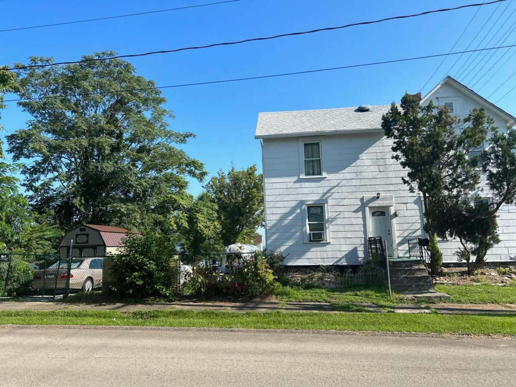 A two-story house with a light-colored exterior is depicted amidst a well-maintained yard. The front entrance features a set of stairs leading to the door, while surrounding greenery adds a natural touch. A parked vehicle is visible in the side parking area.
