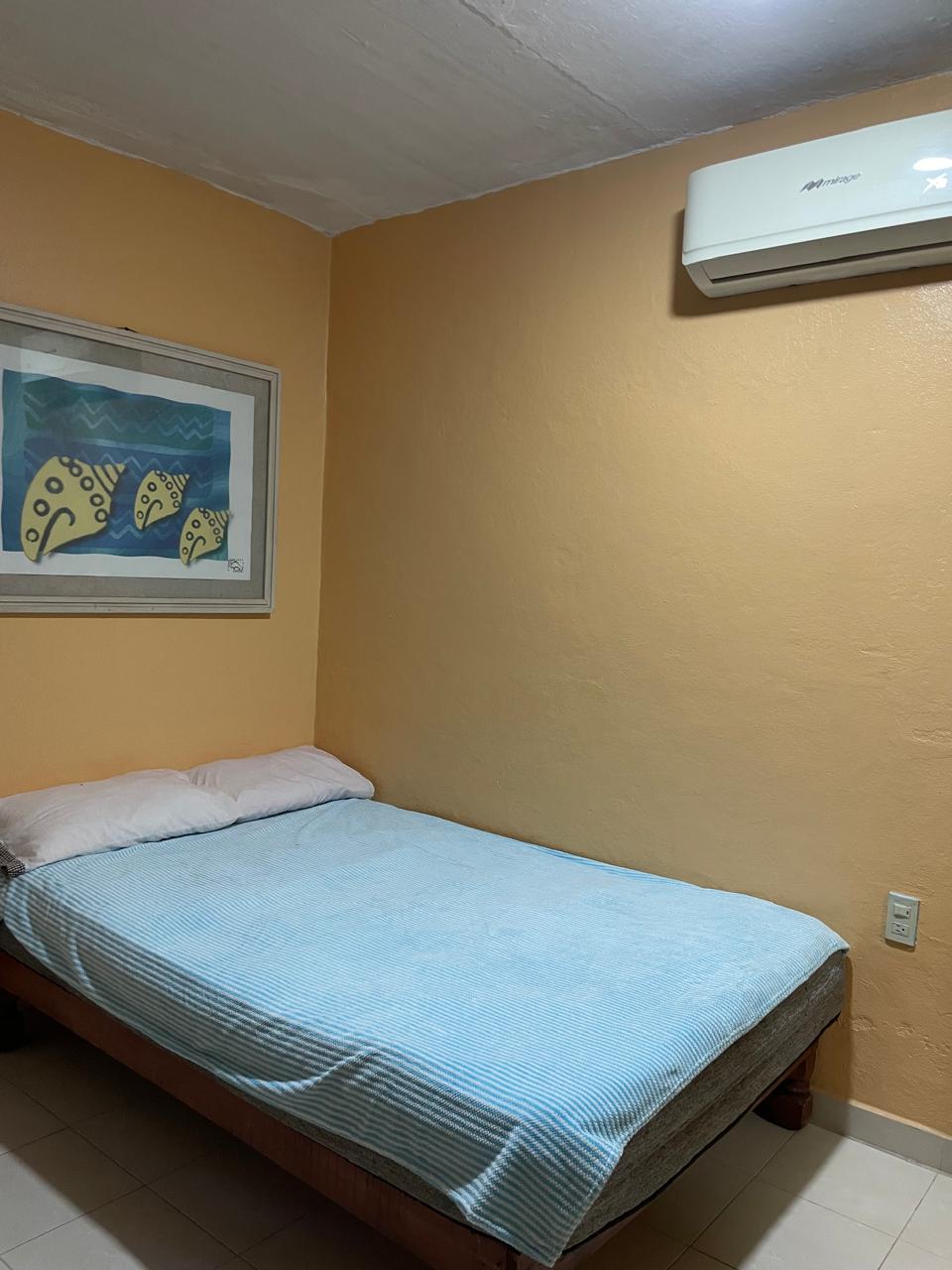 A simple bedroom is equipped with a spacious bed featuring a light blue blanket and two pillows. An air conditioning unit is mounted above the bed, and a framed artwork depicting fish adorns the orange wall. Neutral tiles cover the floor.