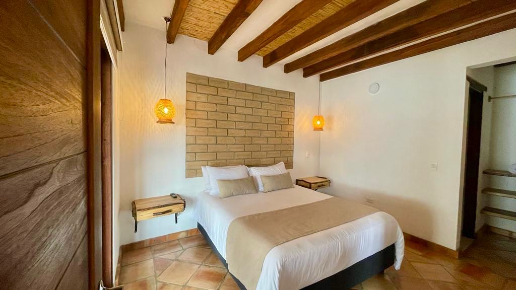 A spacious bedroom features a large bed with neutral bedding against a textured stone wall. Warm, hanging light fixtures provide soft illumination. Wooden bedside tables are positioned on either side, and terracotta tiles enhance the room's earthy aesthetic. Exposed wooden beams complete the inviting design.