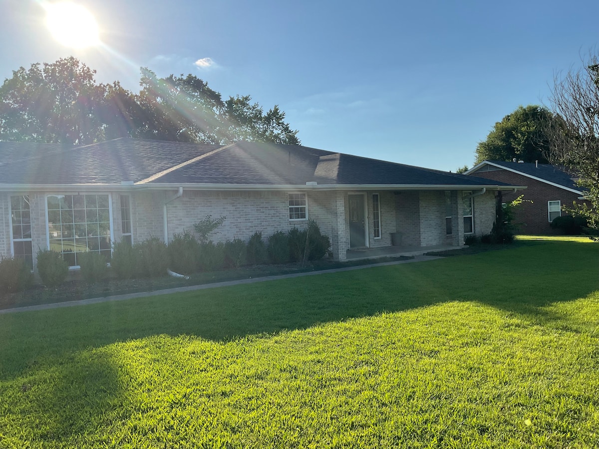 A single-story brick home is captured in the late afternoon light, featuring a well-manicured lawn. Sunlight reflects off the roof, and a neat walkway leads to the entrance, bordered by shrubs and ornamental plants.