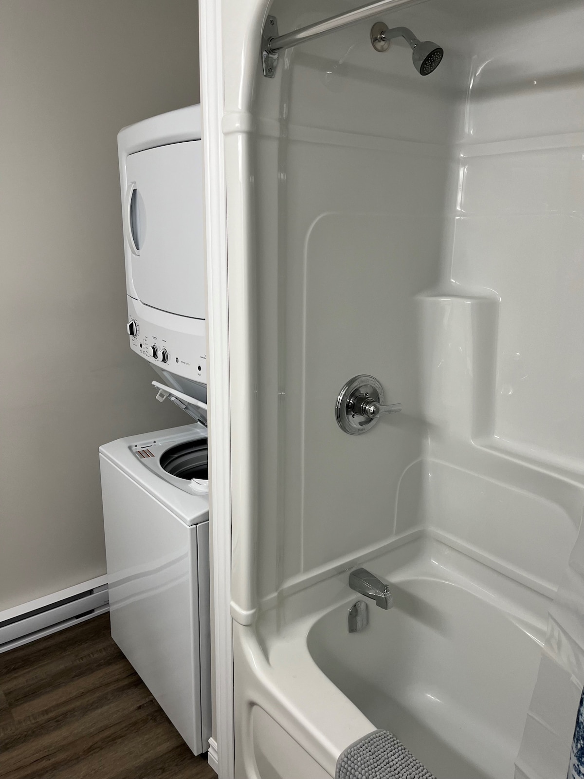 A compact bathroom features a shower-tub combination with a modern faucet. Adjacent to it, a stacked washer and dryer unit provide laundry convenience. The interior is illuminated with neutral-colored walls and a natural wood floor, creating a clean and functional space.
