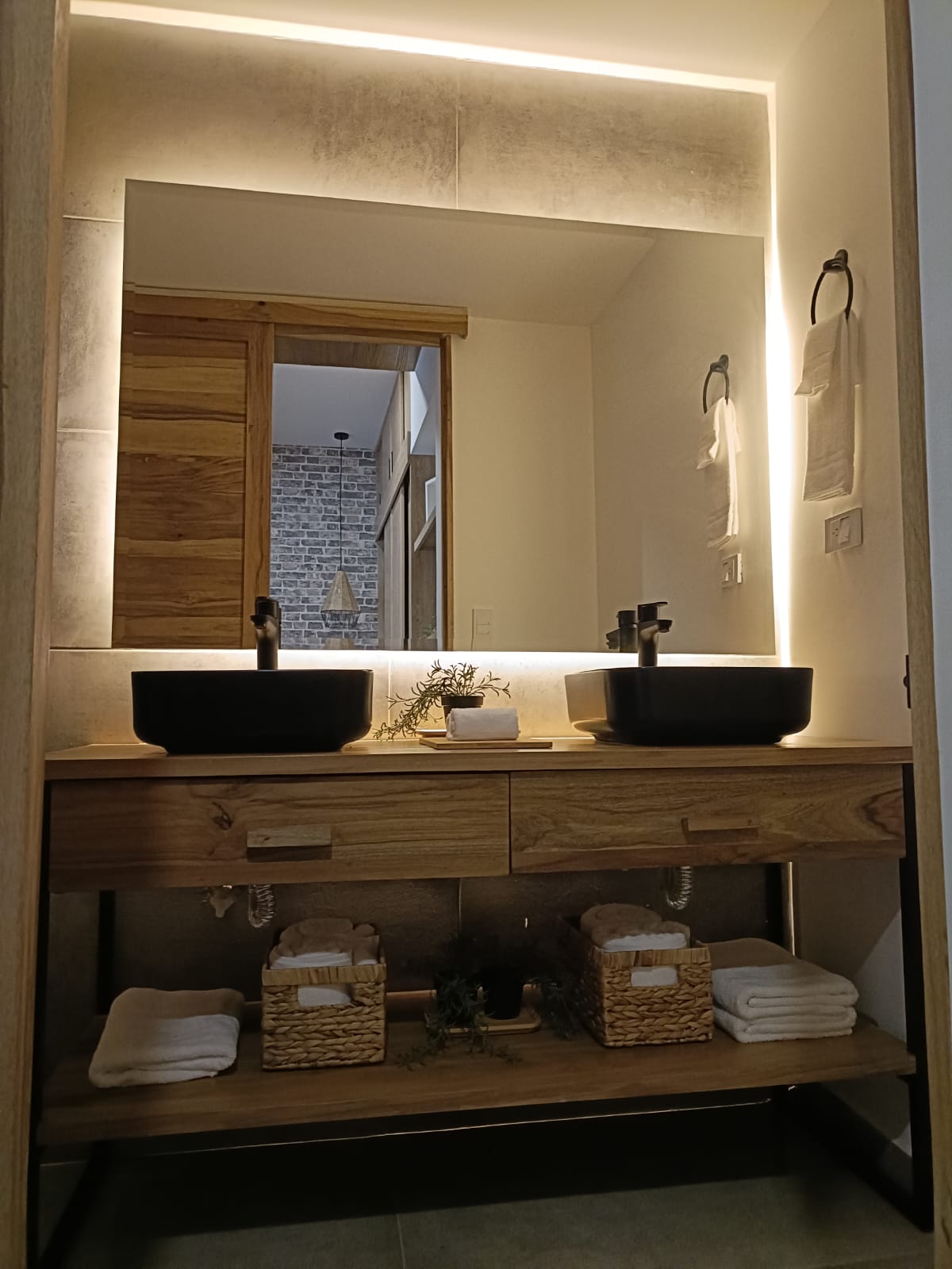 A modern bathroom is featured, showcasing a wooden vanity with two black sinks. Soft lighting frames a mirror above, and neatly arranged towels are visible on a lower shelf. The space is complemented by textured walls and decorative storage bins.