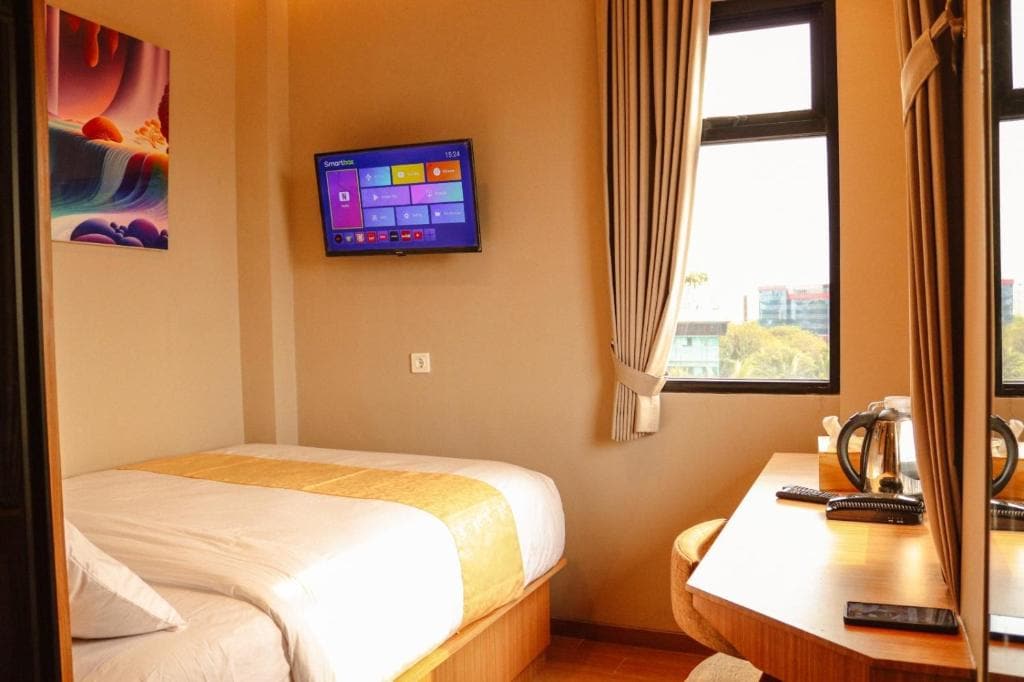 A cozy room features a spacious bed with light linens and a colorful wall art above it. A flat-screen TV is mounted on the wall, while a desk and seating area are visible. Natural light enters through the window, offering a view of the city.