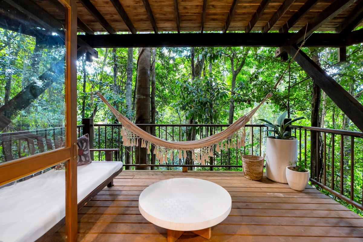 An inviting outdoor space features a hammock suspended between wooden beams, surrounded by lush greenery. The wooden deck showcases a circular table at its center, with potted plants adding touches of nature. Large glass doors connect this serene area to the interior of the treehouse.