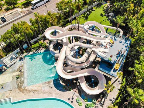 A large water park is depicted, showcasing winding water slides that twist and turn. A spacious pool area can be seen adjacent to the slides, surrounded by lush palm trees. Guests can be spotted enjoying both the slides and the pool.