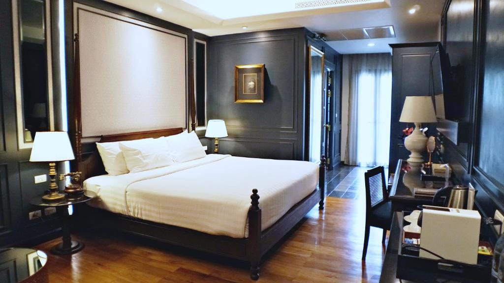 A spacious room is displayed with a large bed centered against dark, textured walls. Two bedside lamps provide soft lighting, and a framed picture adds an artistic touch. A wooden chair and a desk are positioned nearby, while natural light filters through sheer curtains.