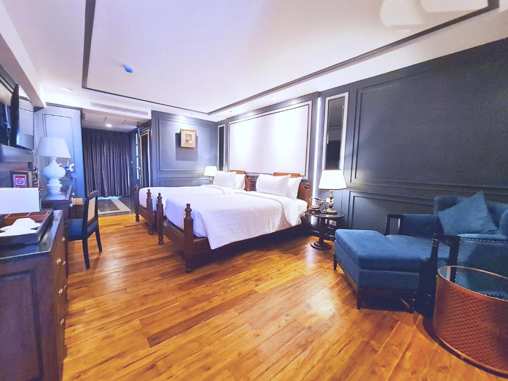 A well-appointed room features a spacious bed with crisp white linens, dark wood furnishings, and a comfortable blue armchair. Warm wooden floors reflect soft lighting, while a desk and a television are positioned along the wall. A curtain-draped window partially filters natural light.
