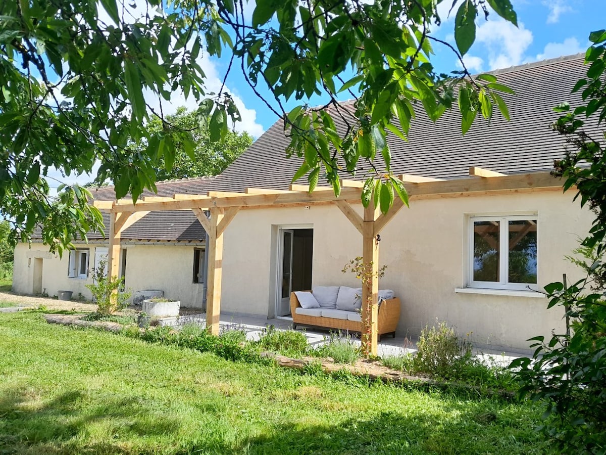 A spacious exterior view of the gîte reveals a covered terrace supported by wooden beams. A comfortable seating area is situated on the patio, surrounded by lush greenery and a manicured lawn. Windows reflect the natural light, enhancing the inviting ambiance.