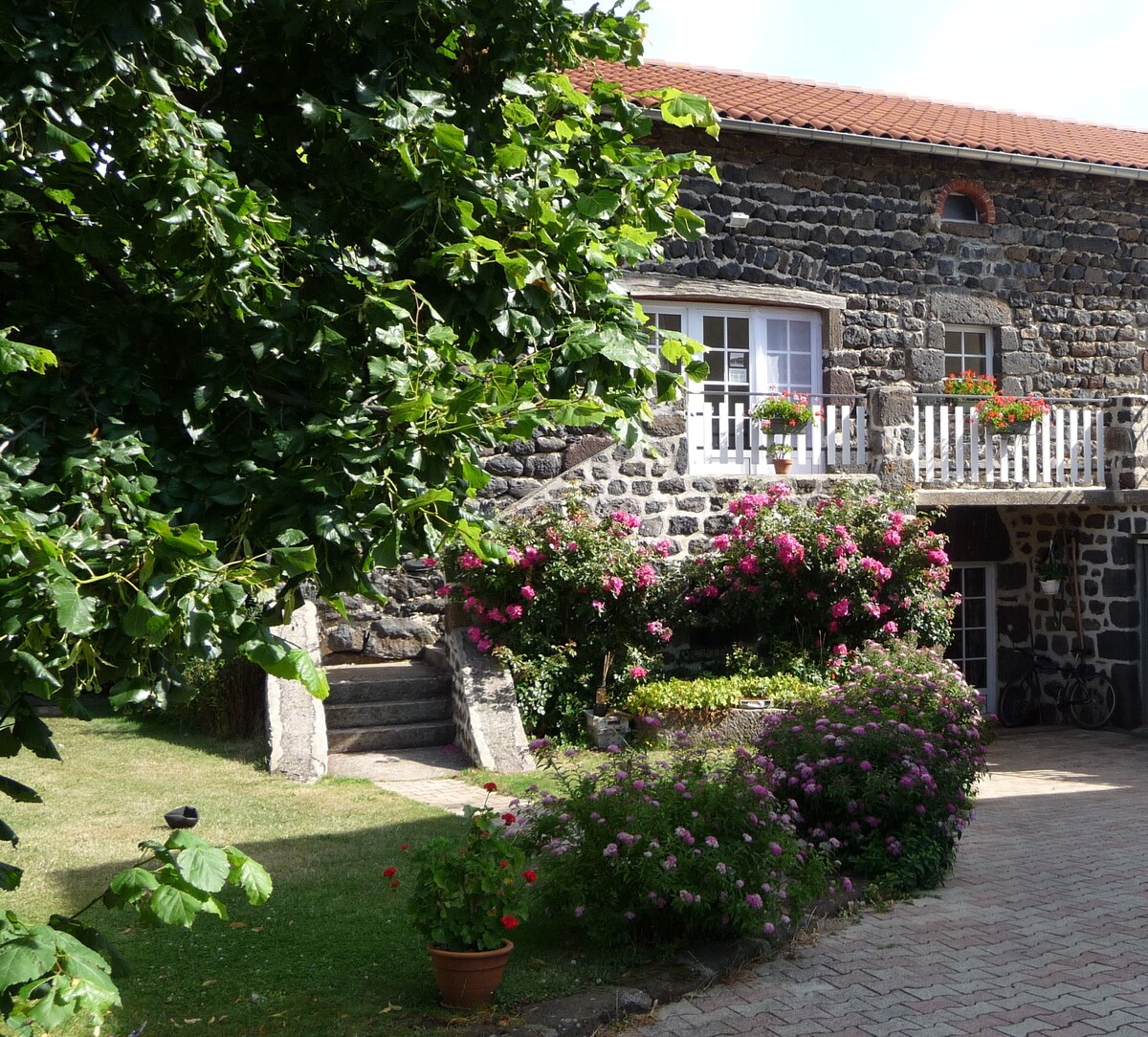 A charming stone exterior of the Gîte d'Amélina is framed by lush greenery and vibrant flowering plants. A staircase leads to the entrance, adorned with window boxes filled with flowers. The pathway is lined with well-maintained landscaping, creating an inviting approach.