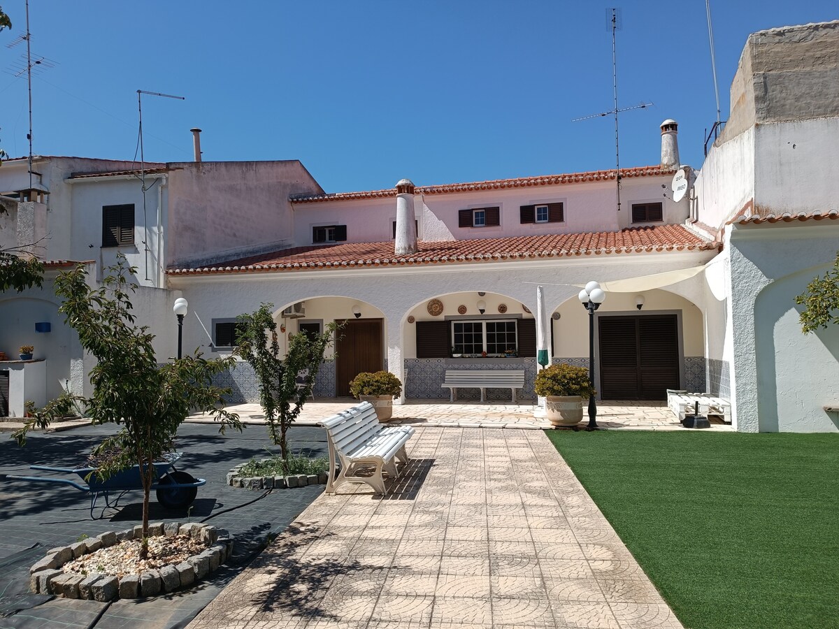 The exterior of the villa is depicted, showcasing a spacious courtyard adorned with green grass and decorative plants. The building features a traditional façade with an arched entryway and multiple windows. Benches are positioned for relaxation, and lighting is provided by lamps along the pathway.