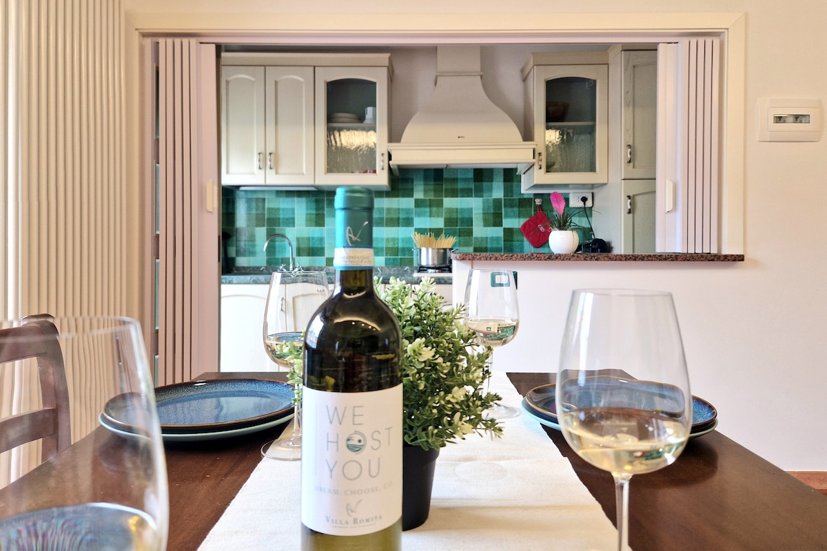 A dining table is set for four, featuring glasses and plates. A bottle of wine is displayed prominently, with a small plant adding a touch of greenery. The kitchen area in the background is visible, showcasing a colorful tile backsplash and cabinetry.