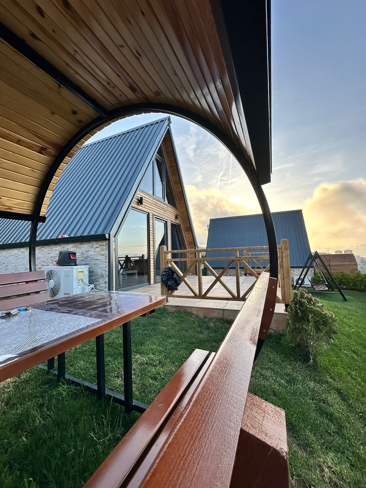 A curved wooden seating arrangement frames the view of multiple triangular-roofed buildings. A spacious deck is visible in the background, with grassy areas accentuating the natural setting. The sky reflects warm hues of sunset, enhancing the inviting outdoor space.