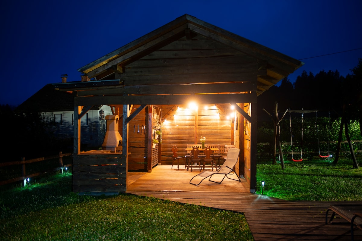 The garden hut features a wooden structure illuminated by warm lights, creating a welcoming atmosphere. A dining table and chairs are arranged beneath the shelter, with two loungers positioned nearby. The surrounding lawn is well-maintained, enhancing the tranquil outdoor setting.
