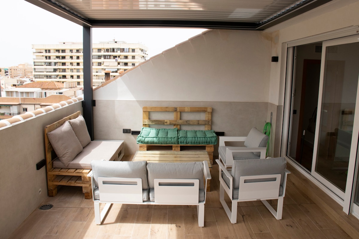 A spacious terrace features a mix of seating options, including a wooden sofa and two cushioned white chairs surrounding a wooden coffee table. A wall partially encloses the space, offering views of the city and ensuring a relaxing outdoor environment.