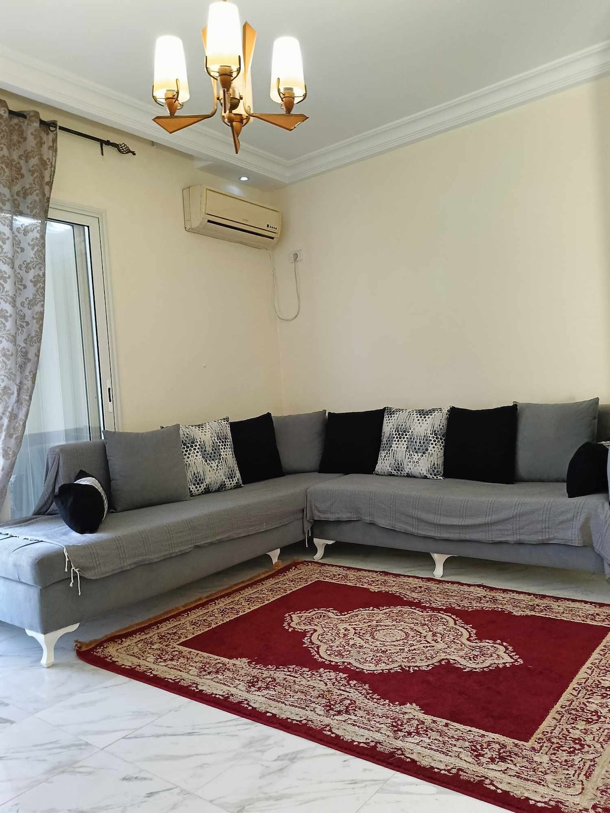 A spacious seating area features a grey sectional sofa adorned with decorative black cushions. A rich red area rug sits beneath, complementing the marble floor. Large windows are dressed with sheer curtains, allowing natural light to fill the room, while an air conditioning unit is positioned above.