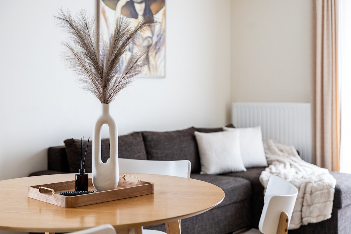 A round wooden dining table is equipped with a beige tray that holds a decorative vase and incense. A cozy sofa with light-colored cushions and a draped blanket is positioned nearby, alongside a wall adorned with artwork. Natural light filters through a curtain-covered window.
