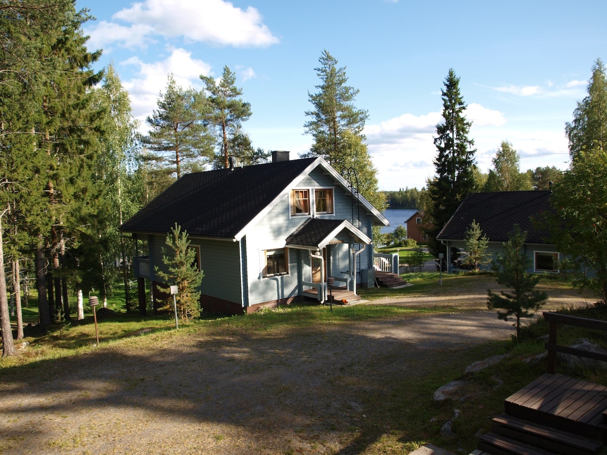 A two-story cabin in a natural setting is surrounded by trees, with a gravel path leading up to its entrance. The exterior features light blue siding and a dark roof. A body of water can be seen in the background, blending with the greenery.