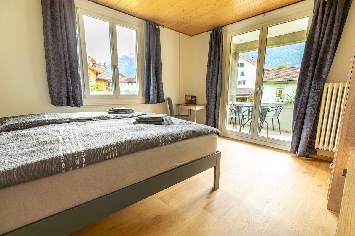 A spacious bedroom features a comfortable bed topped with neat blankets and towels. Large glass doors open to a private terrace, enhancing natural light. The room is equipped with a chair and small table, while curtains frame the windows showcasing views of the surrounding landscape.