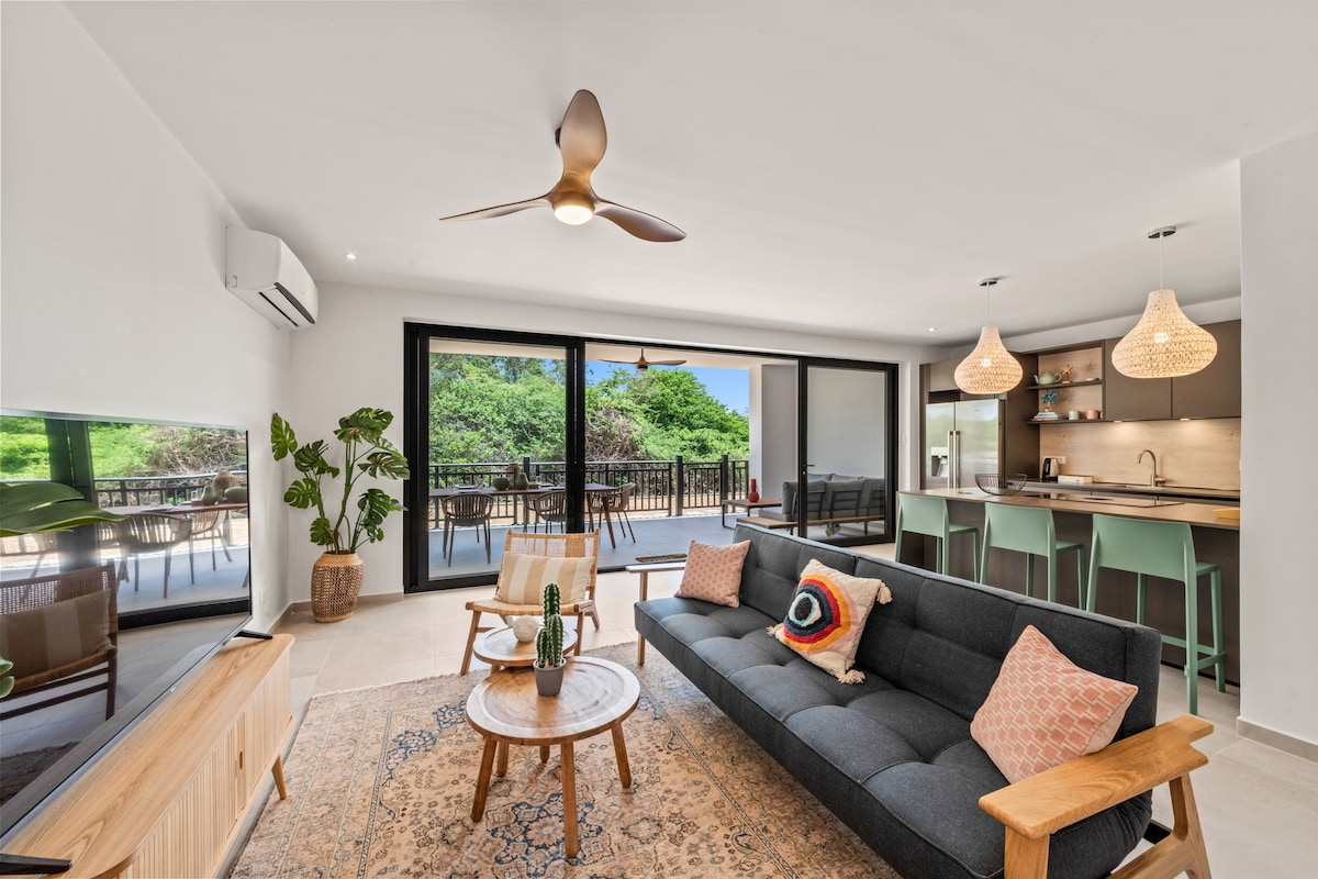 A modern living space features a dark sofa adorned with colorful cushions and a wooden coffee table. Large glass doors open to a spacious terrace surrounded by greenery. A ceiling fan circulates air above, and the kitchen is visible in the background, equipped with contemporary bar stools.
