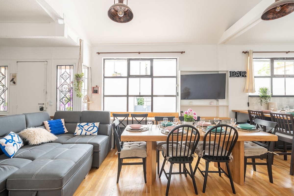 A modern, open-plan living and dining area includes a spacious gray sectional sofa with decorative pillows. A wooden dining table, set for a meal, contrasts with black chairs. Large windows allow ample natural light, and a flat-screen TV is mounted on the wall nearby.