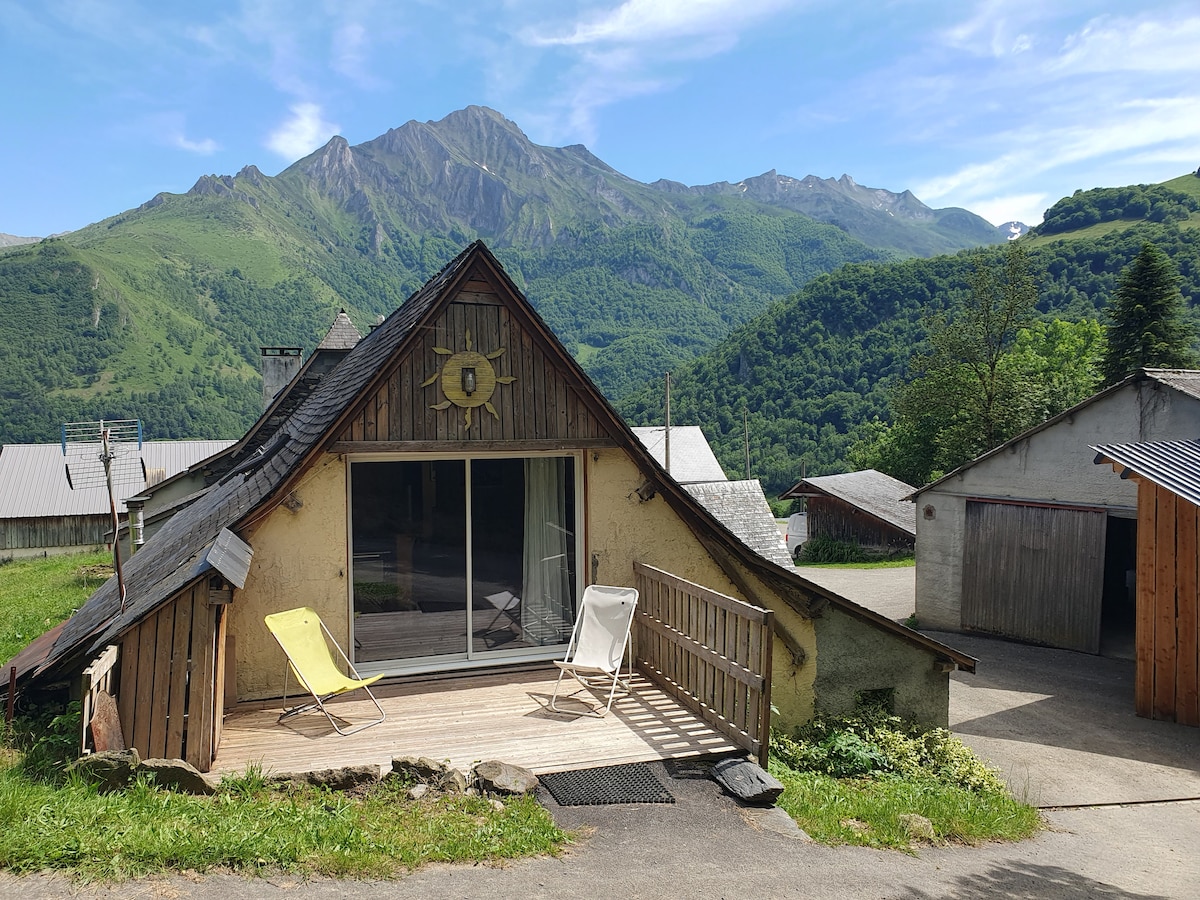A quaint house with a sloping roof is positioned in a serene setting. A wooden deck hosts two chairs, inviting relaxation. Lush green mountains rise in the background, providing a stunning natural view that complements the tranquil surroundings.