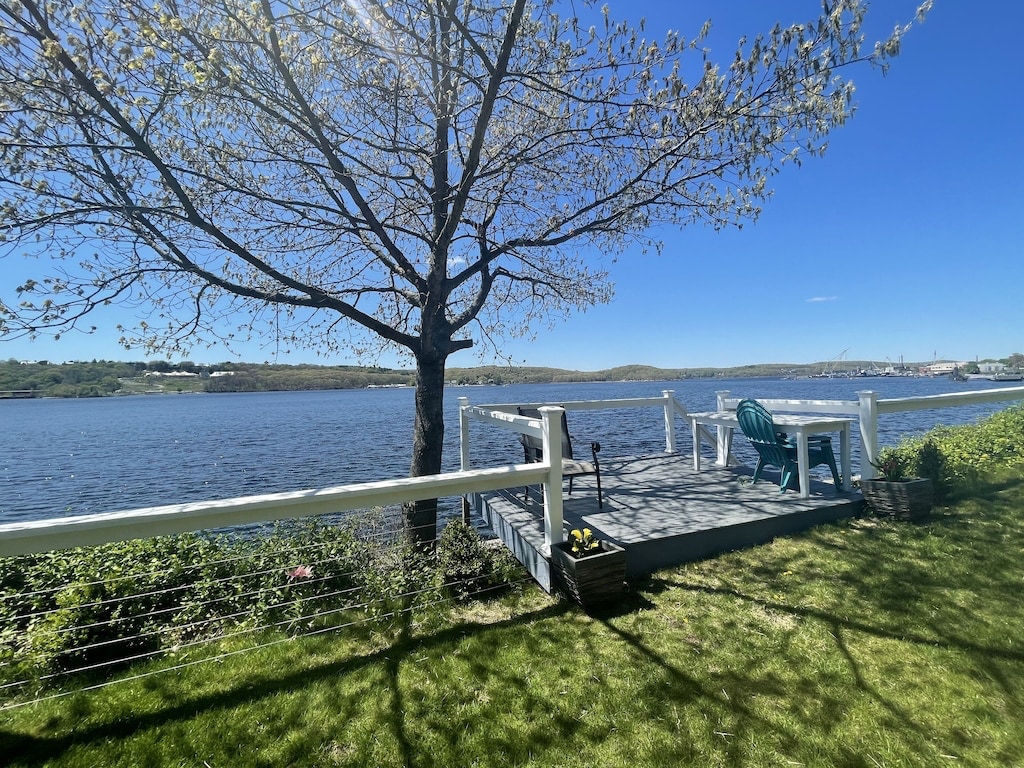 A tranquil outdoor terrace offers views of the waterfront and surrounding landscape. A table and a lounge chair are positioned on the deck, along with greenery along the edge. A tree provides light shade, enhancing the space's inviting atmosphere.