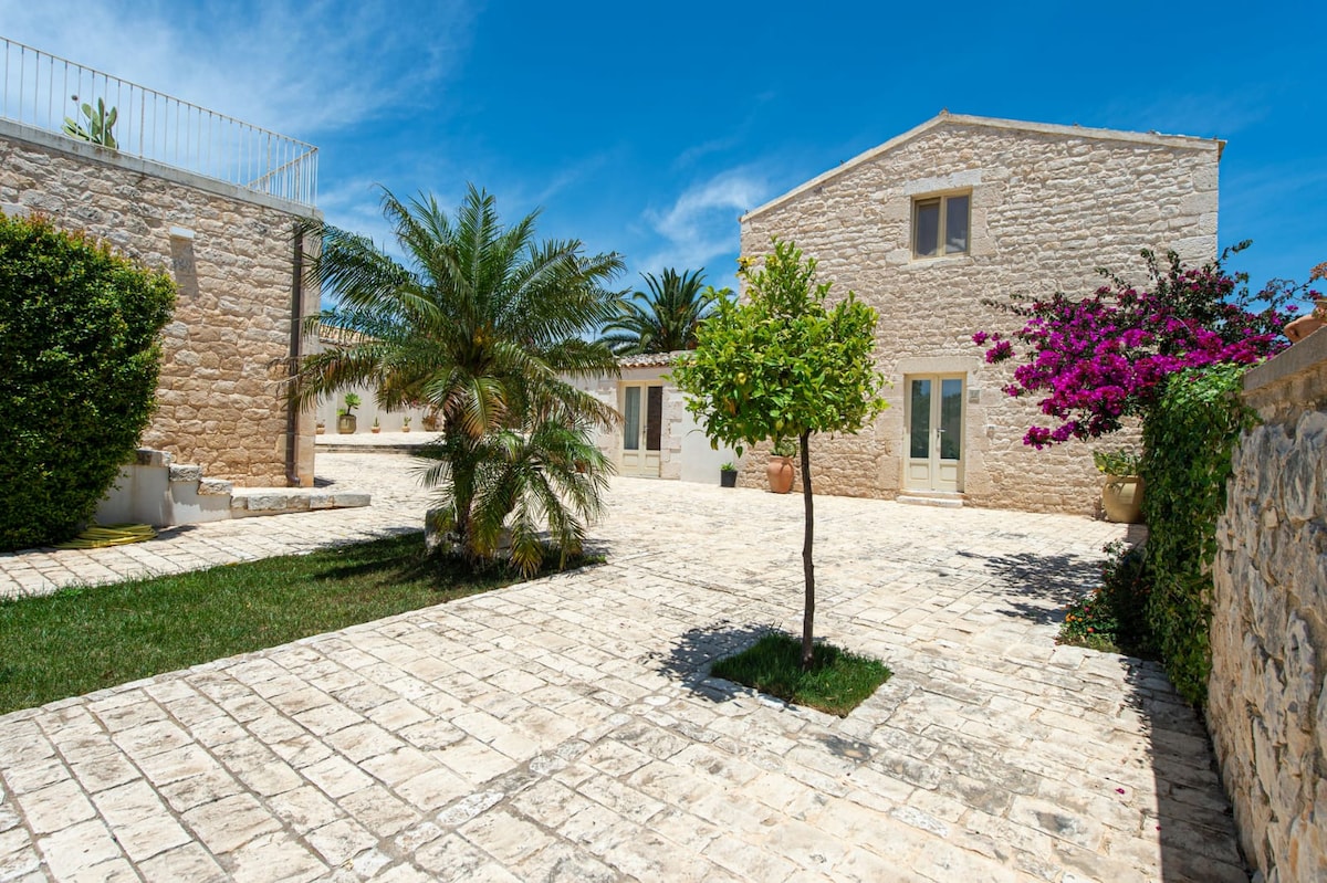 A charming courtyard is framed by natural stone buildings and vibrant greenery. Palm trees and a flowering bush add color to the landscape, while a smooth stone pathway leads to the inviting entrances of the structures under a clear blue sky.