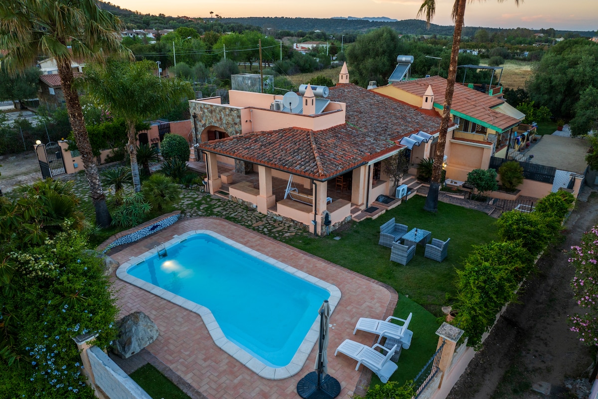 An aerial view reveals a charming villa featuring a private pool surrounded by a patio. Lush greenery and palm trees frame the space, while comfortable seating arrangements are positioned nearby. The terracotta roof and warm tones of the villa are complemented by the tranquil setting.