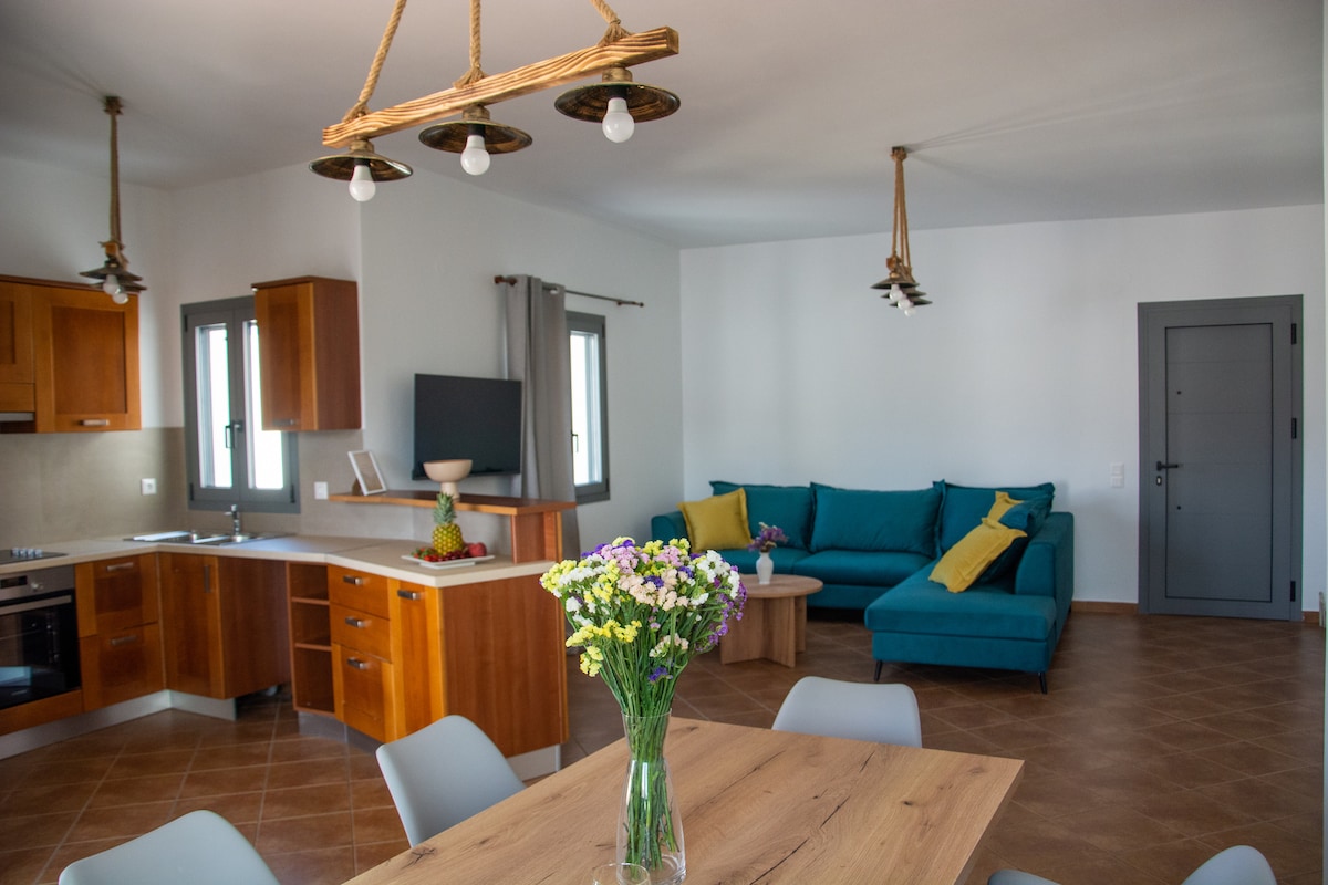 An open-plan living space features a comfortable seating area with a turquoise sofa and yellow accent pillows. A wooden dining table is set beneath a modern light fixture, complemented by a vase of fresh flowers. Natural light floods the room through multiple windows.