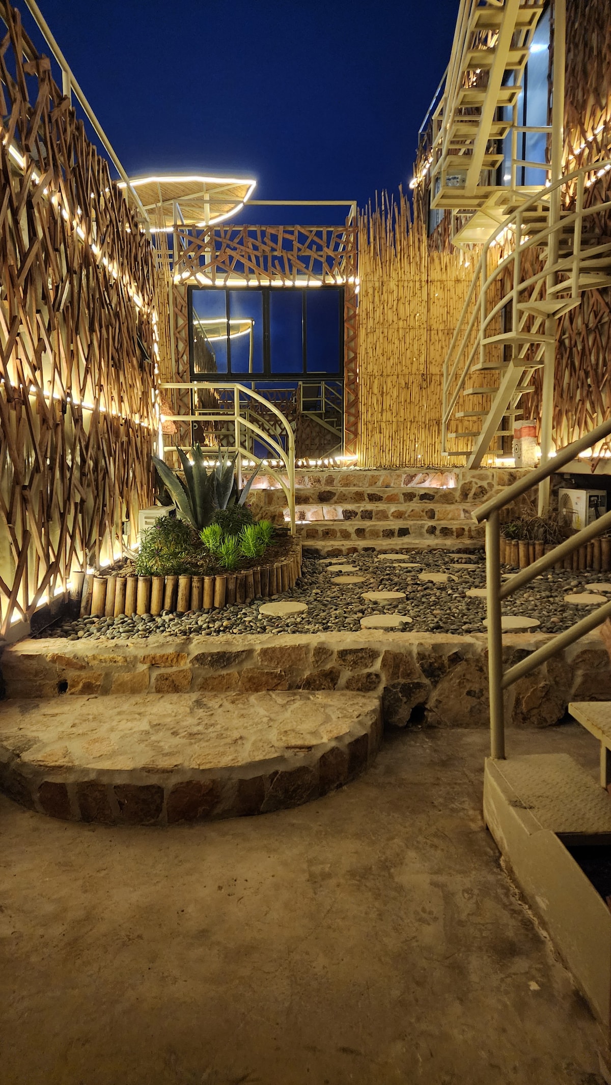 An inviting outdoor pathway is lined with natural stone steps and decorative pebbles, leading towards a unique architectural structure. Soft lighting accents the bamboo features, creating a warm ambiance at dusk, while greenery is strategically placed for added tranquility.