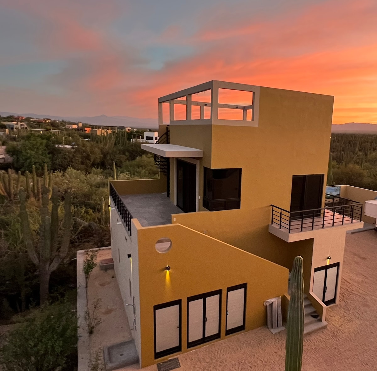 The exterior of Cactus Club Ventana showcases a modern, two-story building against a vibrant sunset sky. A rooftop deck is visible, with a glass railing providing an unobstructed view of the surrounding landscape featuring cacti and distant mountains.